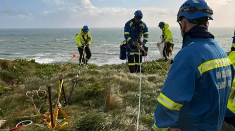 Kingsbridge Coastguard Rescue Team A coastguard rescue training exercise takes place on a grassy clifftop overlooking the sea, with volunteers using ropes, anchors and safety equipment near the cliff edge. Several coastguard rescue officers in high-visibility clothing and helmets can be seen practising rope rescue techniques, while a team member wearing HM Coastguard-branded clothing supervises the operation.