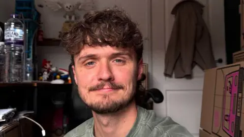 A selfie photograph of Ben Cole Edwards, who is 30 years old, has brown curly hair, a brown beard, and blue eyes. Behind him, a coat can be seen on the hook on his back door, and a plastic water bottle can be seen on a desk behind him. 