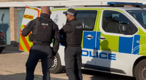 Two police officers standing in front of a marked police van outside a property in Thetford in Norfolk. Both officers have their backs turned to the camera and the van is parked on the road with the back doors of the van wide open.