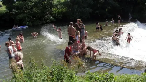 Getty Images Dozens of people of all ages bathe in the water at Farleigh Hungerford. Some sit and stand on the wall of a weir, while others stand waist deep in water. In the background a person can be seen sat in a rubber dingy. 