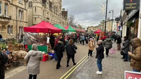 A view of a Christmas market  on Broad Street in Oxford City Centre. Gazebos can be seen erected in the middle of a pedestrianised road. The gazebos' roofs are green and red, and crowds can be seen lining the street with Oxford's historic buildings in the background