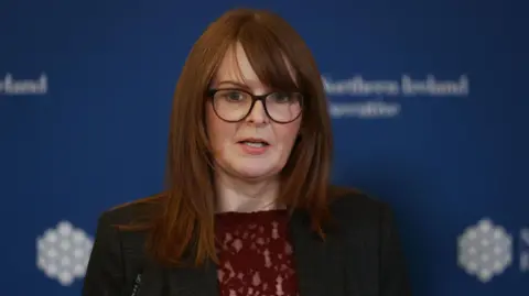 Caoimhe Archibald wearing a dark coloured suit with a blue background behind her. 