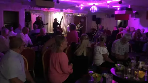Alan Baldwin A live band performs on a small stage inside a community hall at Penton Park, with coloured lights and a disco ball above. Rows of people sit at tables with drinks, facing the stage, in a dimly lit room decorated with bunting and a sign reading “Penton Community and Social Centre”.