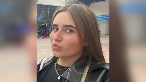 Emily Knell The image shows a woman pursing her lips for the camera. A building can be seen behind her with multiple people walking in and out.