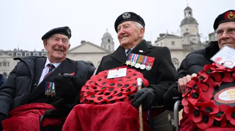 Reuters Stan Ford sitting in the middle of two other veterans during a Remembrance event. They are wearing Navy caps and dark suits, adorned with colourful wartime medals. They are all smiling, with red fluffy blankets resting over their knees and a wreath of poppies on their laps.