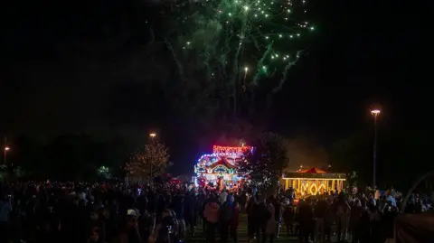 City of Wolverhampton Council Green fireworks in the sky in the distance above crowds of people, above funfair rides at last year's Diwali Mela celebration.