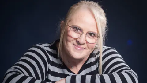 Sarah Templeton A head-and-shoulders picture of Sarah Templeton. She has blond hair tied back, with a few strands left out and framing her face. She has brown eyes and wears round glasses with a thin metal frame. She is leaning on something that is out of shot with her arms folded. She is wearing a striped black and grey top and a silver necklace. She is smiling at the camera.