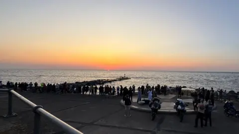People stood looking out to sea from the UK's most easterly point in Lowestoft, with some motorcyclists in the foreground