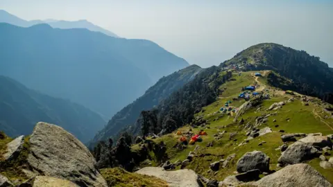 A grass-topped mountain, with some small tents dotted along, with mountains of the Himalayas in the background