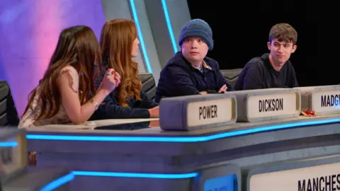 BBC/ITV Studios A side view of four people - a woman with long dark hair and a woman with long red hair, both turned away from the camera, and a person in a blue beanie and a man with short brown hair turned towards them, all sitting behind a TV quiz show desk. In front of them are the words POWER, DICKSON, MADGWICK and MANCHESTER