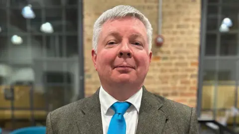 Peterborough City Council Andrew O'Neil looking into camera in the council chamber. He has short white hair and is wearing a grey suit, white shirt and blue tie.