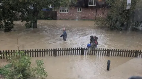 Ness Rodgers Three people wade through high flood waters in Debenham following Storm Babet in 2023. The water reaches their waists and they wear coats. A fence can be seen protruding slightly above the water and homes can be seen in the background. 