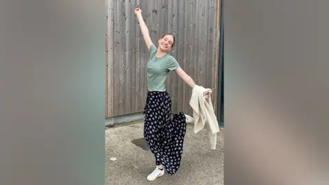 Supplied photo Eleanor stands in a courtyard setting in front of a timber-panelled wall. She's doing a celebration pose, one arm stretched out high above her head and the other pointing towards the ground. One white-trainered foot is off the ground, cocked up behind her. She's wearing a green t-shirt and long, black skirt with a white symbol pattern. She's got her head slightly tilted and is smiling widely.
