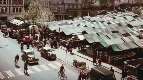 Getty Images Photo shows Norwich Market between 1955 and 1965. There are many stalls with striped canvas canopies, and in the foreground several cars, bicycles and pedestrians are travelling past.