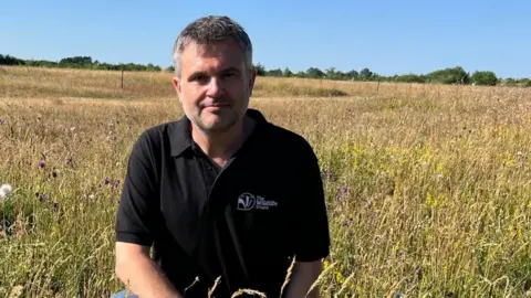 Colin Mackay/BBC Craig Bennett is kneeling in the longs grasses at Trumpington Meadows. Behind him the field is a mix of brown and green with some wild flowers. The sky is a bright blue with no clouds. He is wearing a black top with branding for the Wildlife Trusts written on the left breast pocket. He has short hair, a mix of brown and grey, and some facial hair.