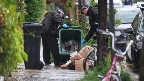 PA Media Police officers search a green recycling bin in Kentish Town, north London.