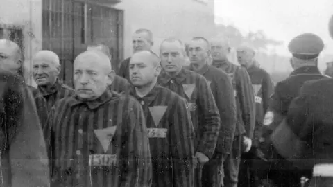 Getty A black‑and‑white historical photograph shows a line of people wearing striped uniforms marked with triangular badges, standing closely together outdoors. The individuals are arranged in a queue beside a building, with uniformed guards visible nearby overseeing the line. The setting, clothing, and badges indicate a controlled detention environment from a wartime period.
