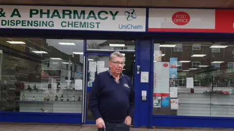 A man in a blue V neck jumper standing in front of the pharmacy and Post Office units which are closing down