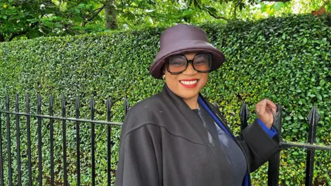 Woman wearing a sunglasses and hat, smiling against a backdrop of green bushes and trees.