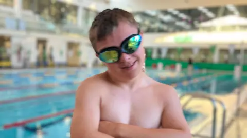 Sam Craddock wearing reflective swim goggles stands beside an indoor swimming pool with arms crossed. He is positioned in the foreground near the pool edge. The background shows swimming lanes, poolside railings, and a spacious aquatic center interior with bright lighting and blurred architectural details.