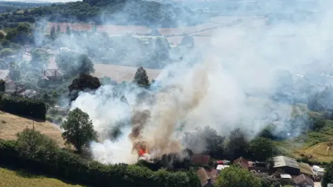 Devon and Somerset Fire and Rescue Service Aerial shot of a blaze by a farm. There is some open grassland in the background and a large plume of white smoke.