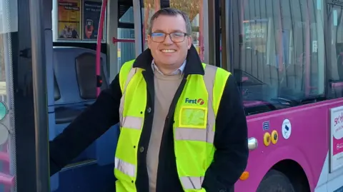 First Bus A man stands next to a bus, with his right hand leaning on the handle of the open door. He is wearing a hi-vis jacket over a navy coat, a beige jumper and a white shirt. He has short hair and dark-framed glasses.