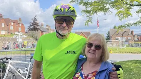 Nicky Patrick / BBC A man in a fluorescent yellow top with a cycling helmet on. He is stood next to a woman and has his arm round her shoulder.