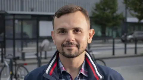 Dalibor Sentic Dalibor Sentic is photographed stood outside on a street. A bike, bus stop and trees can be seen behind him. He has brown eyes, short brown hair, and a beard. He is wearing a plaid grey, red and white shirt and navy-blue jacket, which has a white and red stripe on the collar.