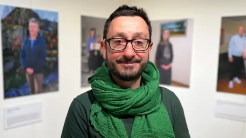 Photographer Aaron Yeandle stood in front of four of his portraits. He is wearing a dark green jumper with a green scarf.