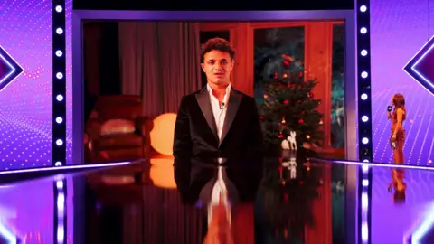 Lando Norris addresses the Sports Personality of the Year Awards via video link. He is on a screen wearing a suit jacket and in front of a Christmas Tree. The shot is of the screen on a purple lit stage with a shiny floor and the presenter looking at him to the right of the shot.