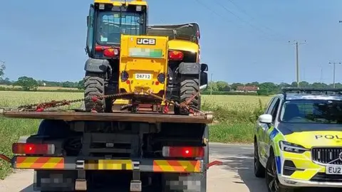 Essex Police A JCB telehandler and trailer. An Essex Police car which is fluorescent yellow and blue is parked next to it.
