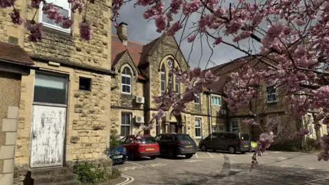 The exterior of the hospital. It is a yellow brick building with Victorian features. Cars are parked in bays outside the front of the hospital, with a large blossom tree to the side of it.