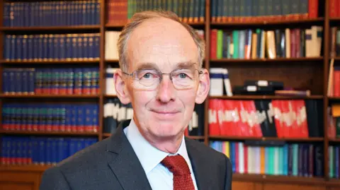 Judicial Office Sir Andrew McFarlane. He is wearing a dark grey suit, light blue shirt and red tie. He is looking at the camera with a wooden bookcase full of books behind him. He has short grey hair and glasses
