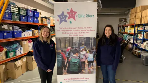 BBC Leanne is blonde and is wearing a dark blue hoodie that says "little stars" on it. She is standing next to a large banner which has the charity logo on it, with pictures of children walking to school, alongside information about the charity. On the right of the banner is Lindsey, a volunteer, who is wearing a dark blue hoodie and has long brown hair