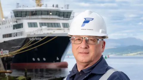 A man with a hard hat, wearing glasses, with a large ship in the background 