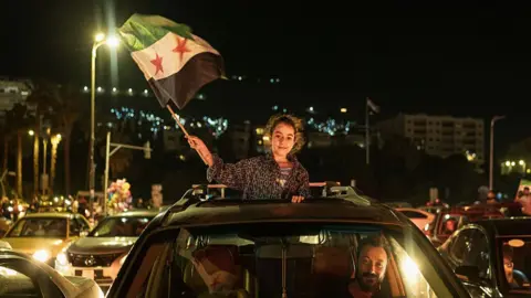A girl waves a Syrian flag out of the open roof of a car being driven by a smiling man on streets filled with traffic in Damascus