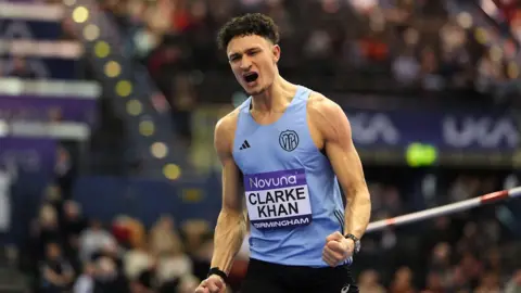 An athlete with black curly hair celebrates while wearing a light blue sports vest with "Clarke Khan" pinned to his shirt. 