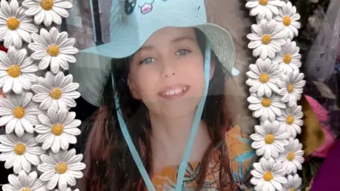 Family handout A picture in a frame. The frame is made up of plastic white daisies. The picture is of a smiling young girl with long brown hair. She is wearing a bright blue sun hat.