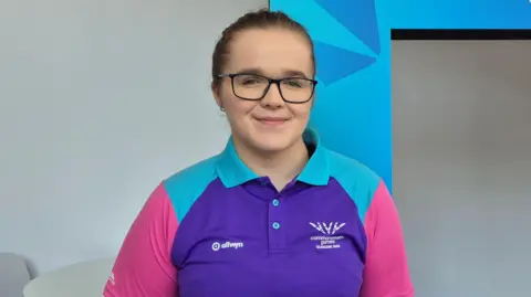 A woman standing in front of an advertising prop for the Commonwealth Games. She is wearing glasses, has short dark hair and is wearing a top with purple, aqua and pink colours.