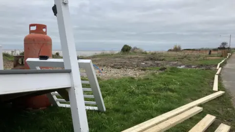 GUY CAMPBELL/BBC In the foreground, a red gas cylinder in standing upright next to discarded white fencing, with an empty, grassy clifftop and cloudy sky in the background 