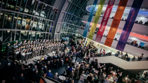 The Glasshouse The concourse of the Glasshouse with a choir singing on the left side. There are two balconies with people looking over them. There are five colourful banners hanging from the ceiling. People are lining the stairs through the centre of the room. The wall behind the choir is made completely from glass and is reflecting the lights from inside the concourse.