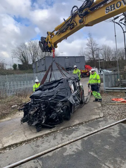 The wreckage of a car being lifted by a crane, surrounded by three men in high-vis clothing.