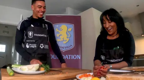 Zena Weeks Zena Weeks (right) and Morgan Rogers are both laughing, with food on the table in front of them and an Aston Villa Foundation sign behind them. They are both wearing black clothing and black aprons that include the word Foundation on them.
