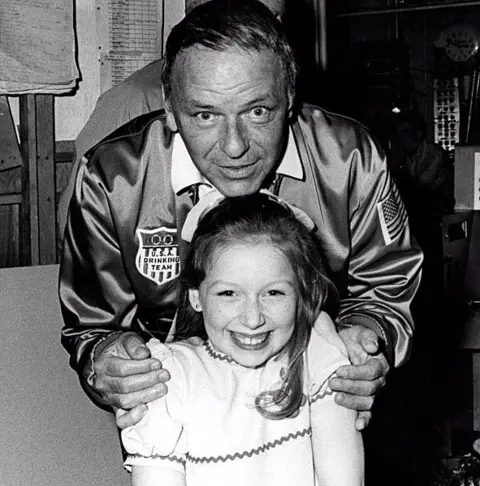 STAX/Specky Productions/BBC Scotland Black and white photo of Frank Sinatra, in a baseball bomber jacket, holding a young Lena Zavaroni's shoulders and leaning over her. Lena is grinning and is wearing a white dress and a big white bow in her hair.