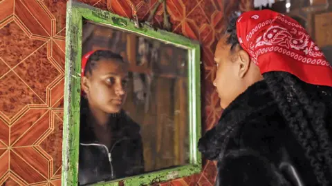 A young mixed race woman looks at herself in a mirror - she is wearing a black zip-up top and has braided long hair