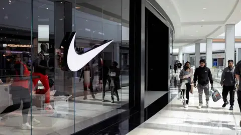 A white Nike swoosh is seen on a store inside a mall with shoppers walking by