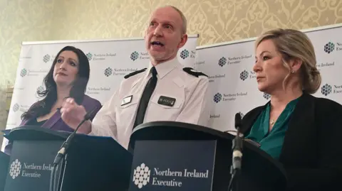 Pacemaker Minister Emma Little Pengelly (wearing a purple jacket), Assistant Chief Con Davy Beck (wearing a PSNI uniform) and Minister Michelle O'Neill (wearing a dark green top) standing at pulpits with signs saying Northern Ireland Assembly. 