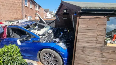A wide shot of the Blue Skoda crashed into the garden shed.