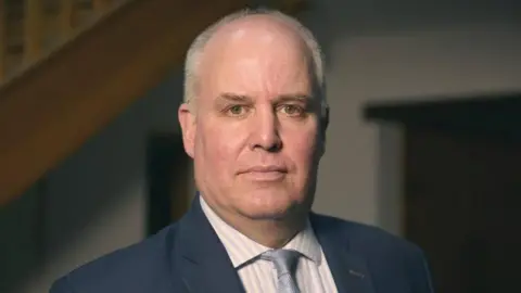 BBC Andrew RT Davies is looking at the camera, wearing a dark blue suit, a stripy white shirt and a light blue tie. He is standing with a staircase in darkness in the background.