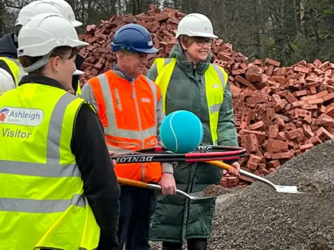 A group of people in hard hats and wearing high visibility vests hold shovels in their hands while one of them has a tennis racquet with a large tennis ball on top of it